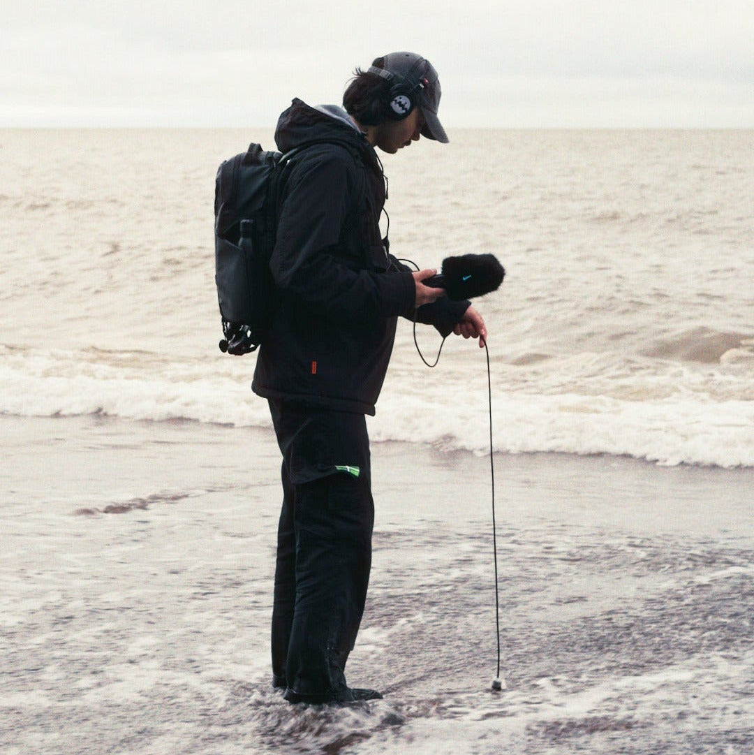 Person with a backpack and audio equipment standing in water on a beach.